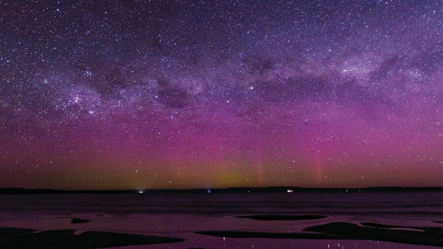 The purple and pink hues of the Aurora Australis in the Tasmania night sky. The stars are seen reflecting in pools of water.